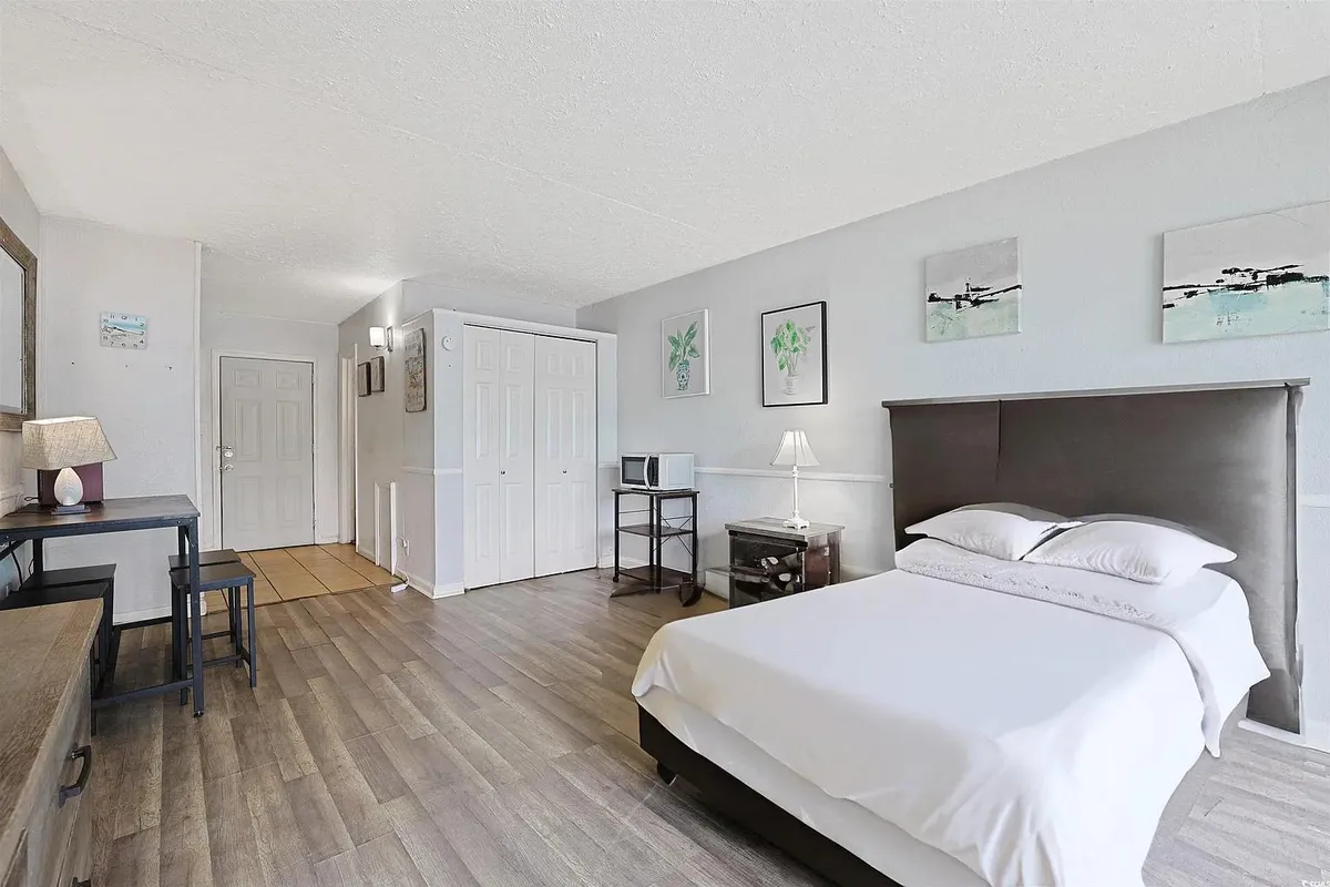 A cozy and thoughtfully arranged studio interior featuring a plush bed, a sleek desk perfect for reading or working, and tasteful artwork. The space feels calm and inviting, with a closet for convenience and natural light streaming in, creating an ideal spot for morning coffee by the window or relaxing after a day out.