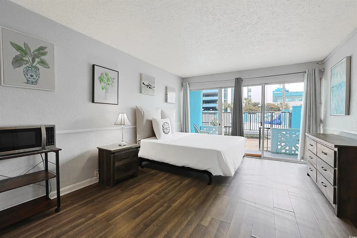 The image features a serene studio space with a cozy bed positioned near a large sliding glass door. Soft natural light streams into the room, illuminating tasteful decorative plants and offering a glimpse of the balcony—perfect for leisurely mornings. The view beyond the door hints at an open, inviting outdoor space, ideal for relaxing or sipping coffee while enjoying Myrtle Beach's gentle atmosphere.