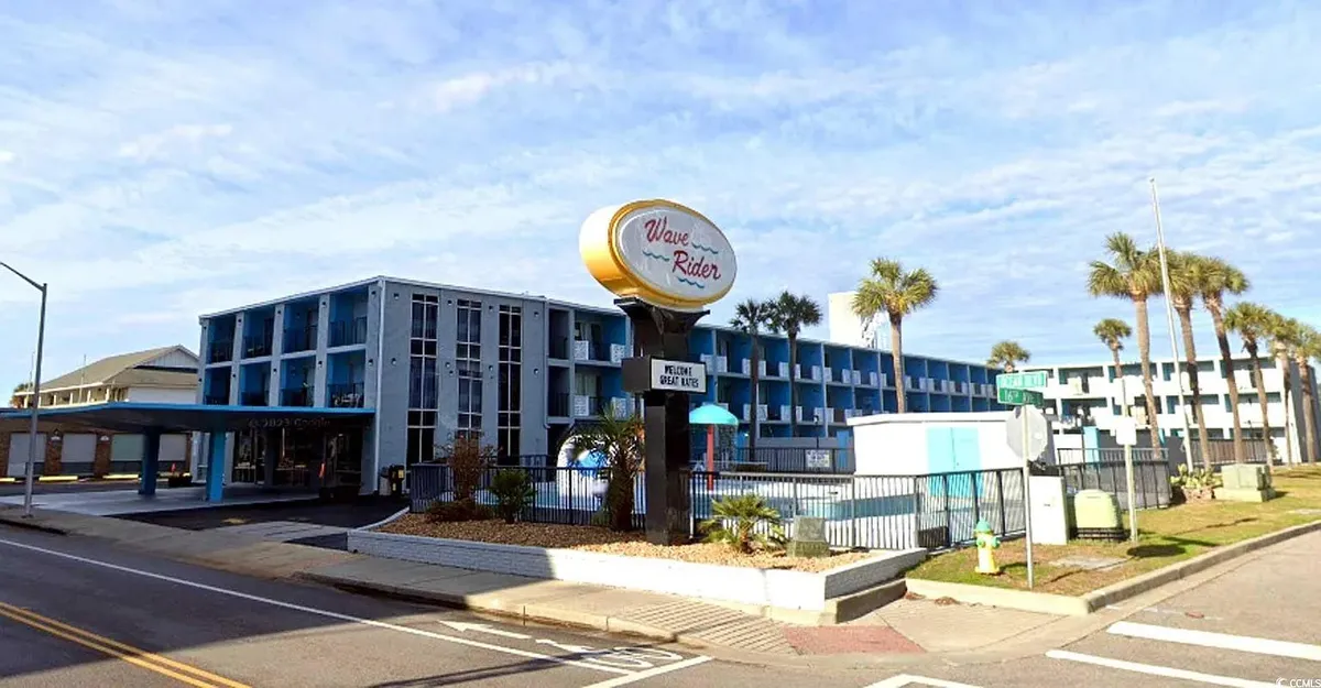 This image presents the main entrance of the resort, flanked by palm trees and featuring the hotel sign and building façade. In the background, guests can be seen swimming and sunbathing near the pool, while others explore the grounds or head toward nearby beach access. The scene is suffused with a relaxed ambiance and hints at easy movement between the pool, hotel, and beach.