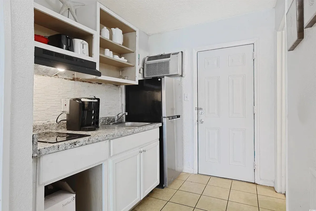 A modern kitchenette equipped with a stylish countertop, sleek black refrigerator, and open shelves displaying white dishware. The space is bright and functional, with a door leading outside—ideal for preparing a morning coffee or enjoying a casual meal in comfort.